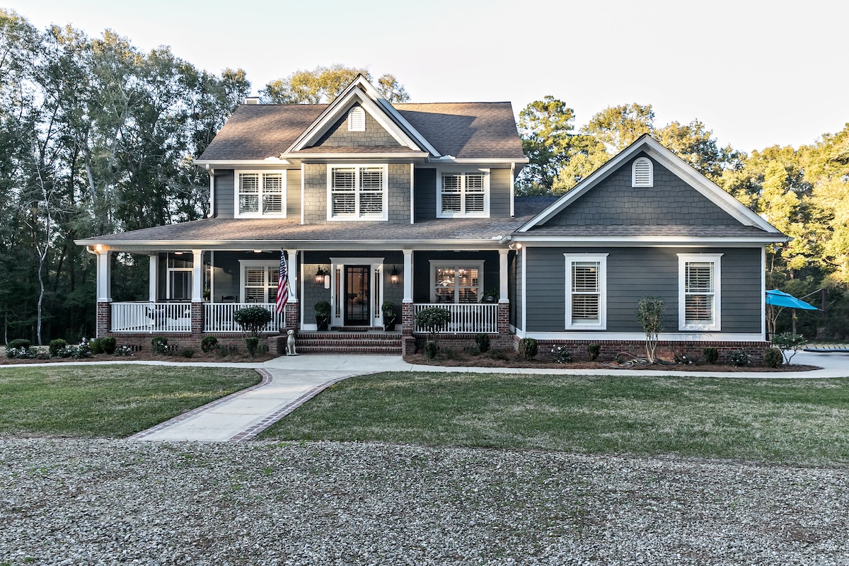 Front view of a large two story blue gray house with wood and vinyl siding with an expansive porch, sidewalk and manicured lawn.