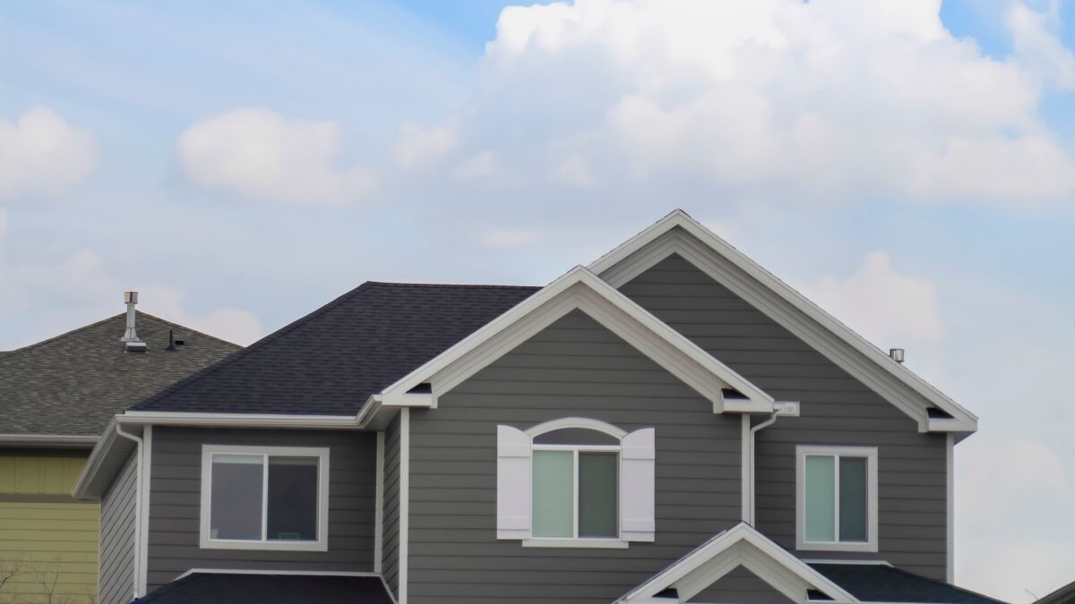 Panorama Exterior of the top floor of a house with blue sky and bright clouds background. The home has a pitched roof, gray wall, and arched window with shutters.