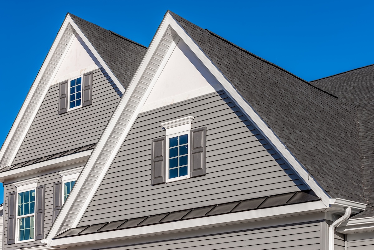 Double gable, with white decorative trim over the windows on a triangle gable roof, white soffit and fascia, gray horizontal vinyl lap siding with blue sky background