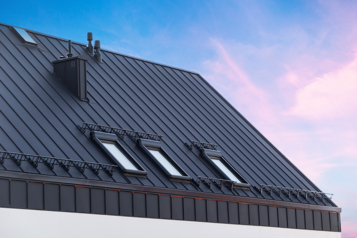 how long do metal roofs last House with metal galvanized coating, chimney pipe, mansard windows and snow holding cornice on roof slope against sunset sky. Element of modern residential building with zinc rooftop sheets