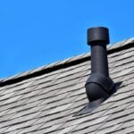 Black air ventilation chimney on grey shingles roof of residental house on blue sky background with copy space for text.